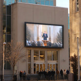 Large outdoor screen displaying a video of a speaker in front of an audience on a building facade.