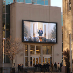 Large outdoor screen displaying a video of a speaker in front of an audience on a building facade.