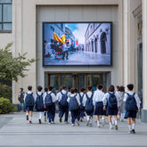 School children walking past a large screen displaying a video, with a building and trees in the background.