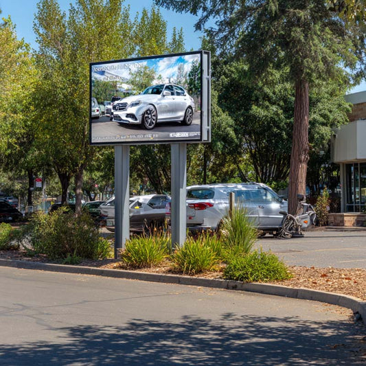 Billboard displaying a car advertisement in an outdoor setting with trees and parked cars.