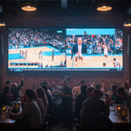 People watching a basketball game on a large screen in a bar.