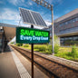 Pole-mounted solar-powered outdoor LED sign with top solar panel displaying Save Water message by railway platform