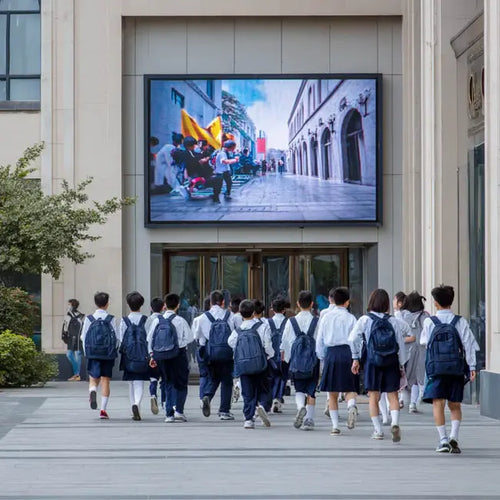 School children in uniforms and backpacks walking past a large screen displaying an urban scene.